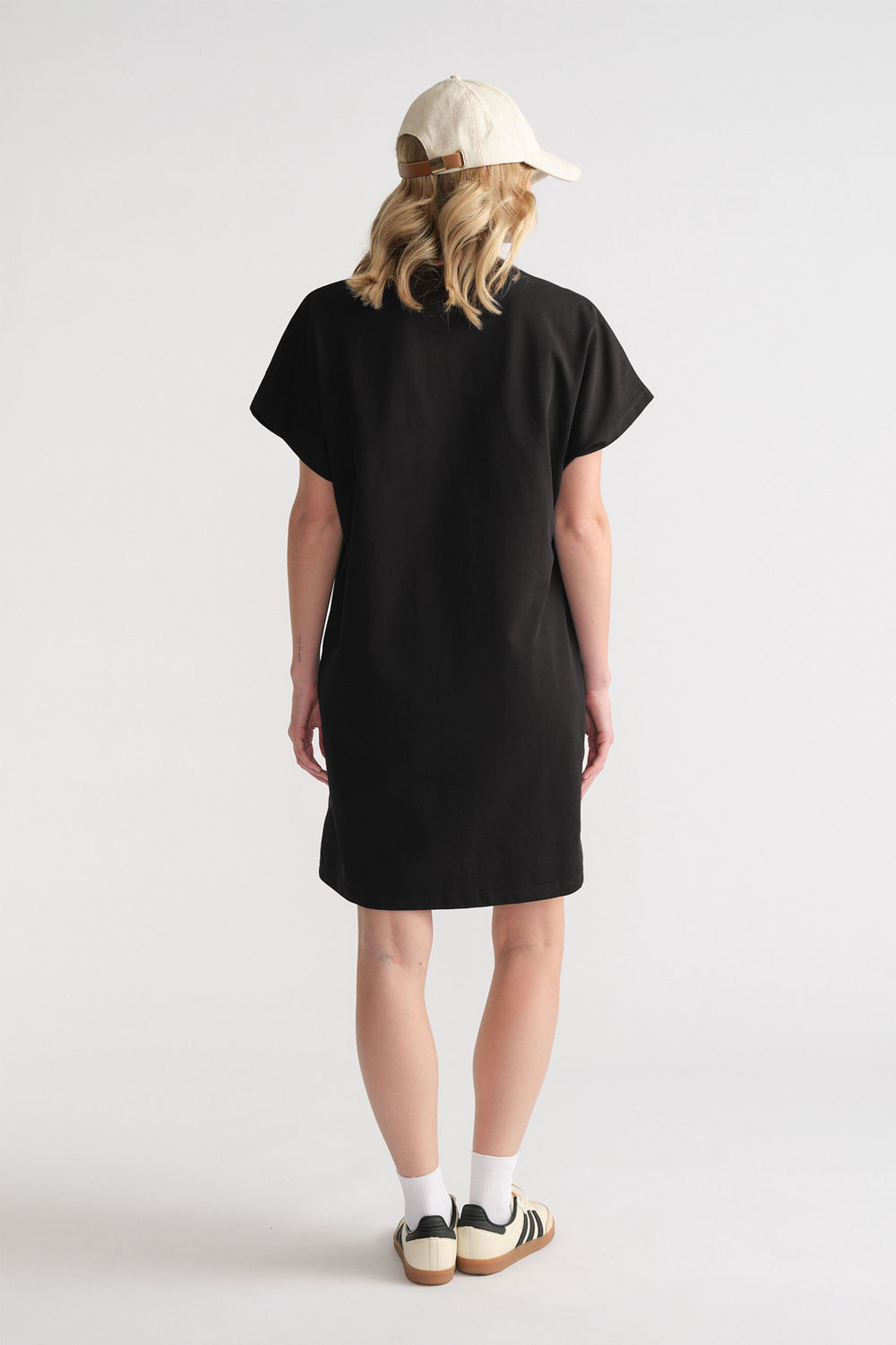 Back view of woman wearing a relaxed black short tee dress paired with white socks, sneakers, and a light beige cap