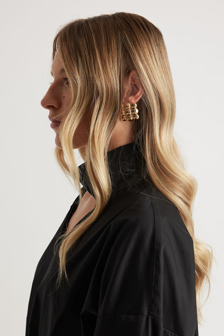 Side profile of woman with long wavy blonde hair wearing chunky gold statement stud earrings and black collared top