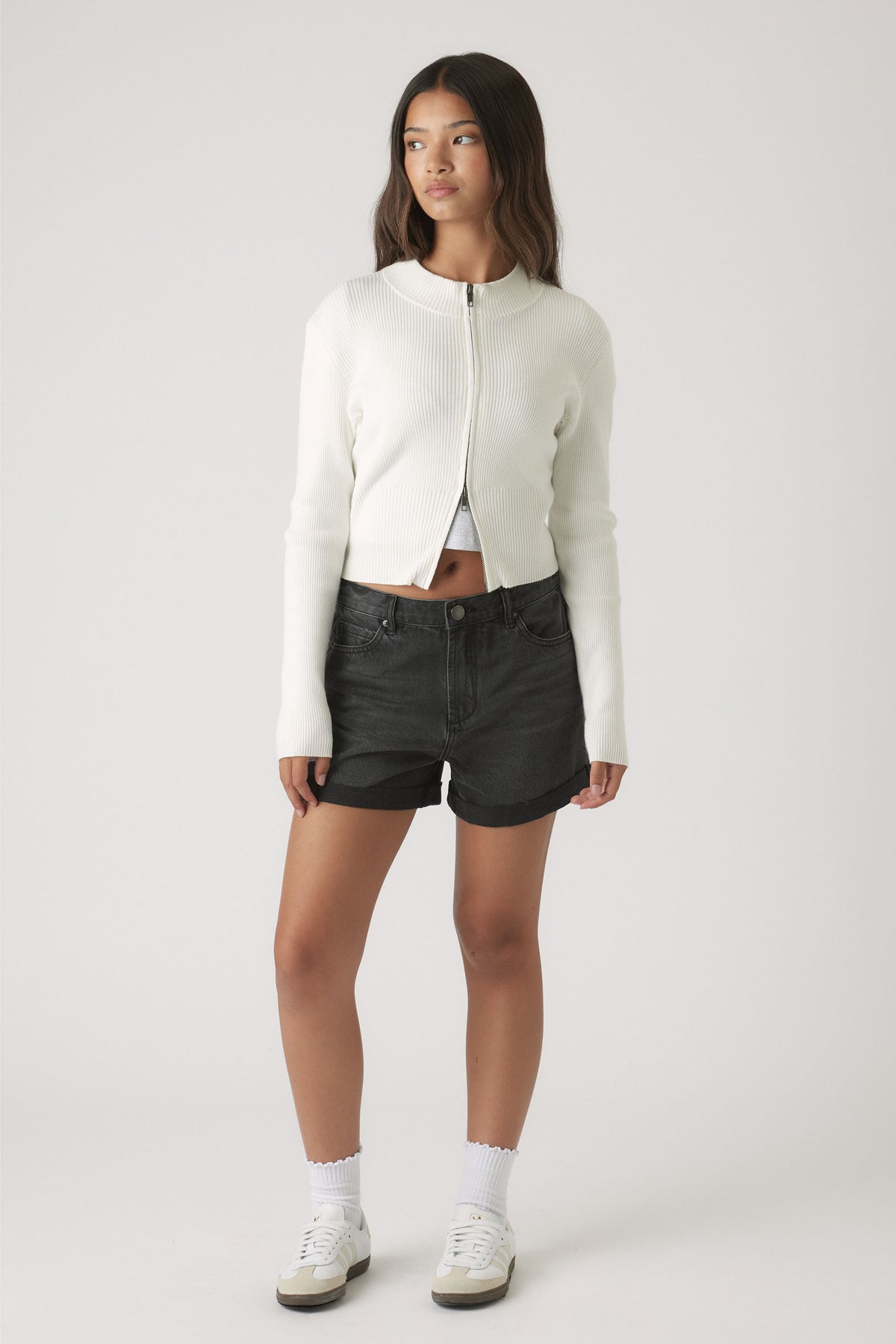 Teen girl wearing a cropped white zip-up sweater and black denim cuffed shorts for casual, effortless style.