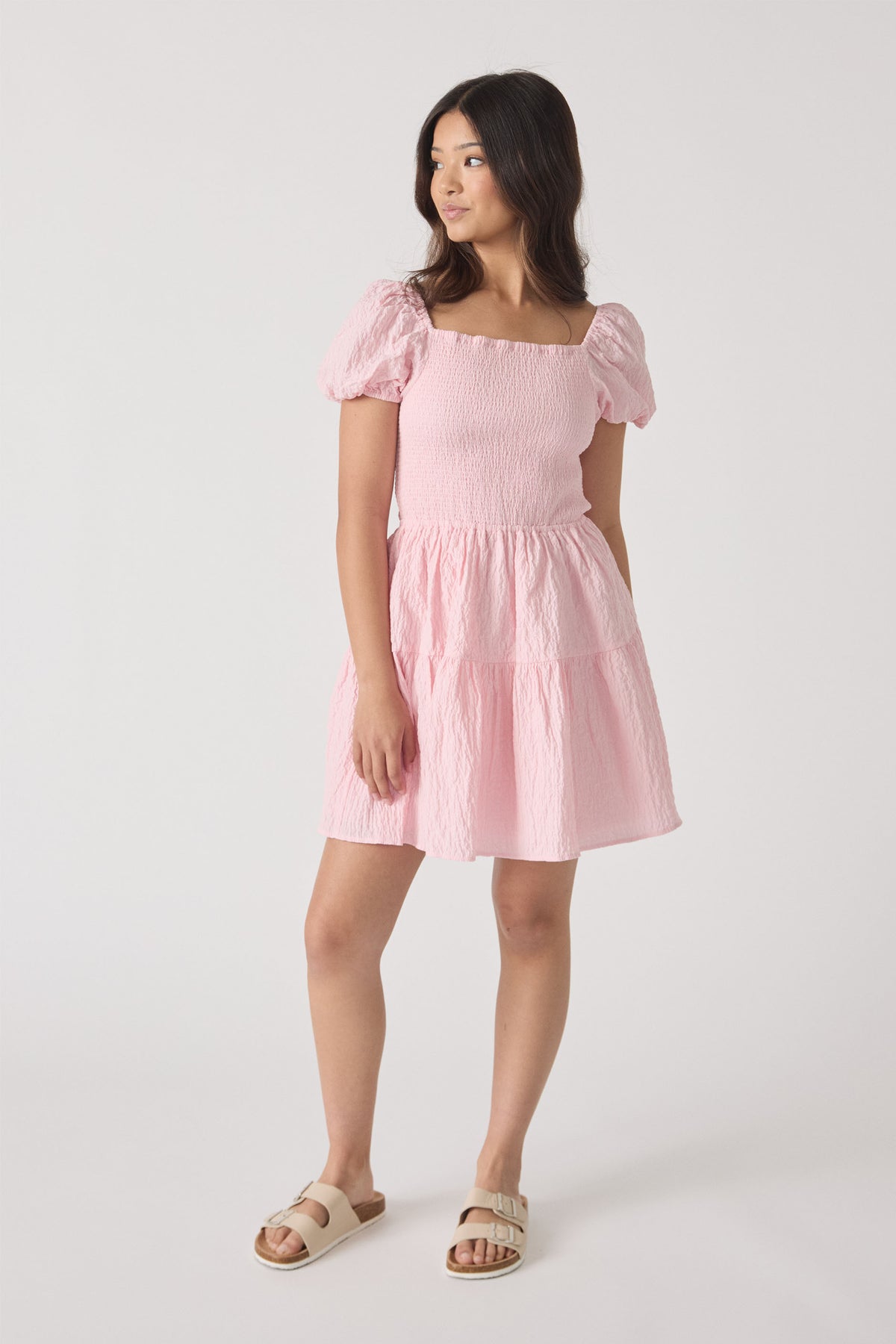 Girl wearing a soft pink mini dress with puff sleeves and smocked bodice, paired with beige buckle sandals.