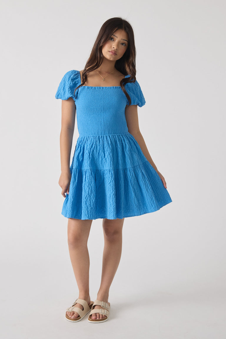 Teen girl wearing a bright blue textured mini dress with puff sleeves and square neckline, paired with beige sandals.
