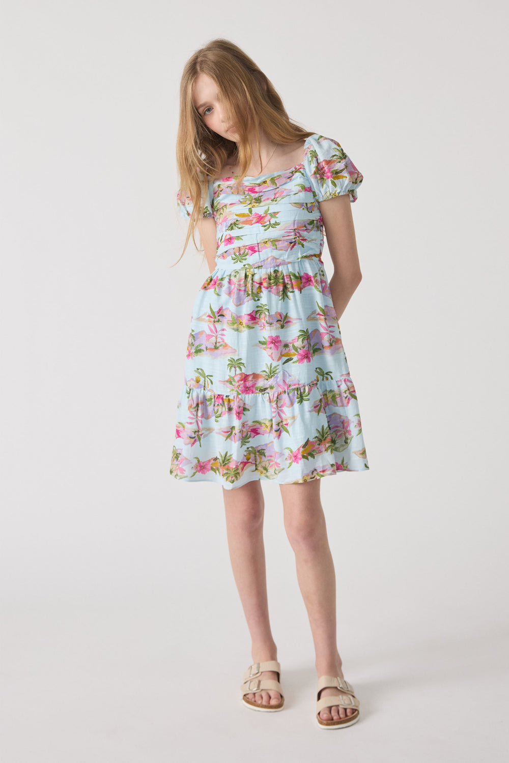 Girl wearing a light blue mini dress with puff sleeves and a tropical floral print, paired with beige sandals.