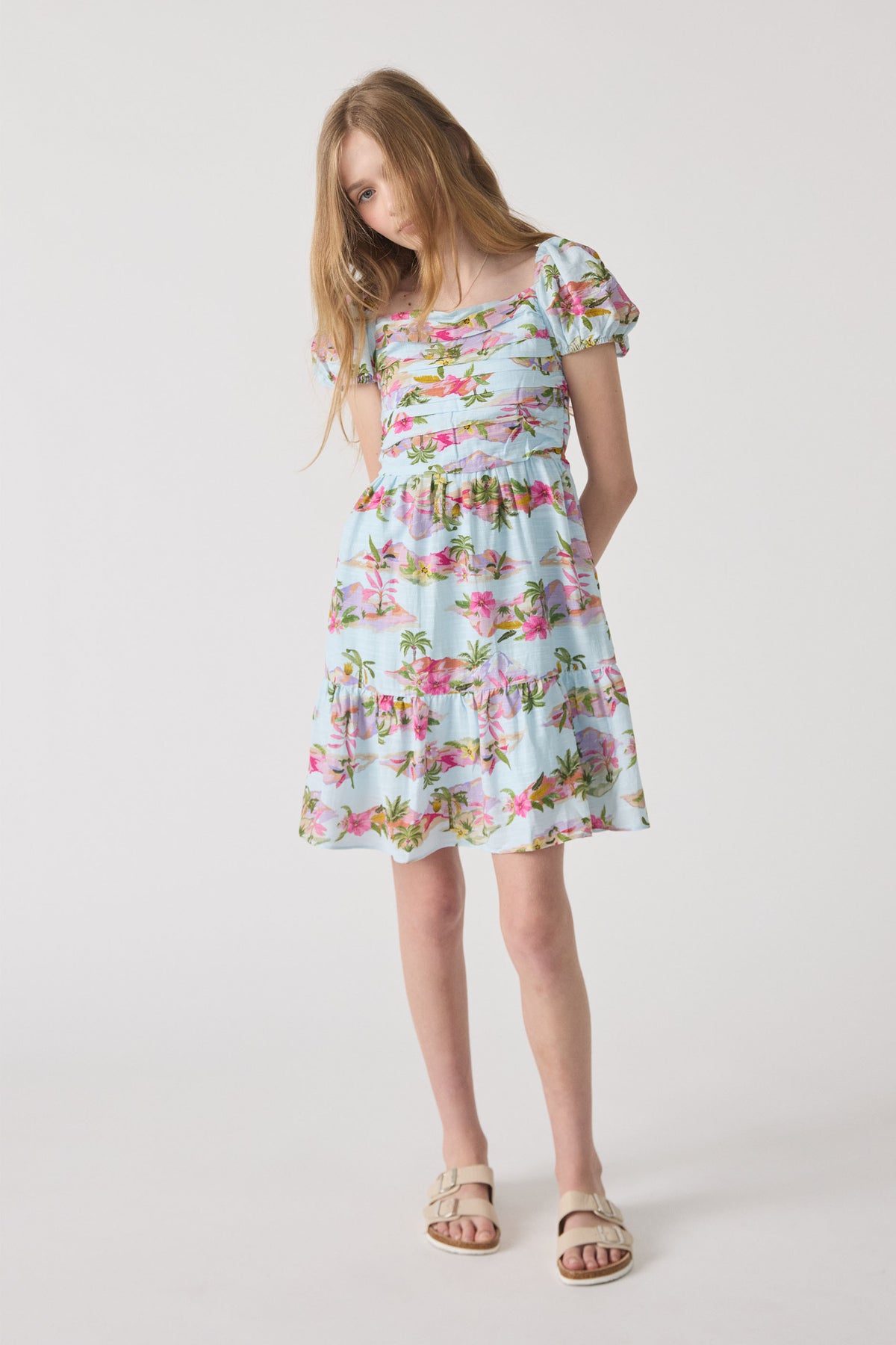 Girl wearing a light blue mini dress with puff sleeves and a tropical floral print, paired with beige sandals.