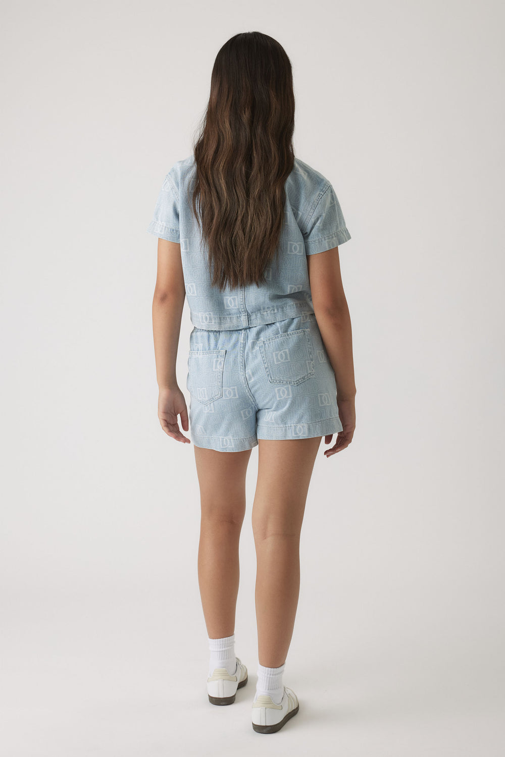 Girl wearing light blue denim shorts and matching short-sleeve shirt with subtle monogram print, paired with white sneakers.