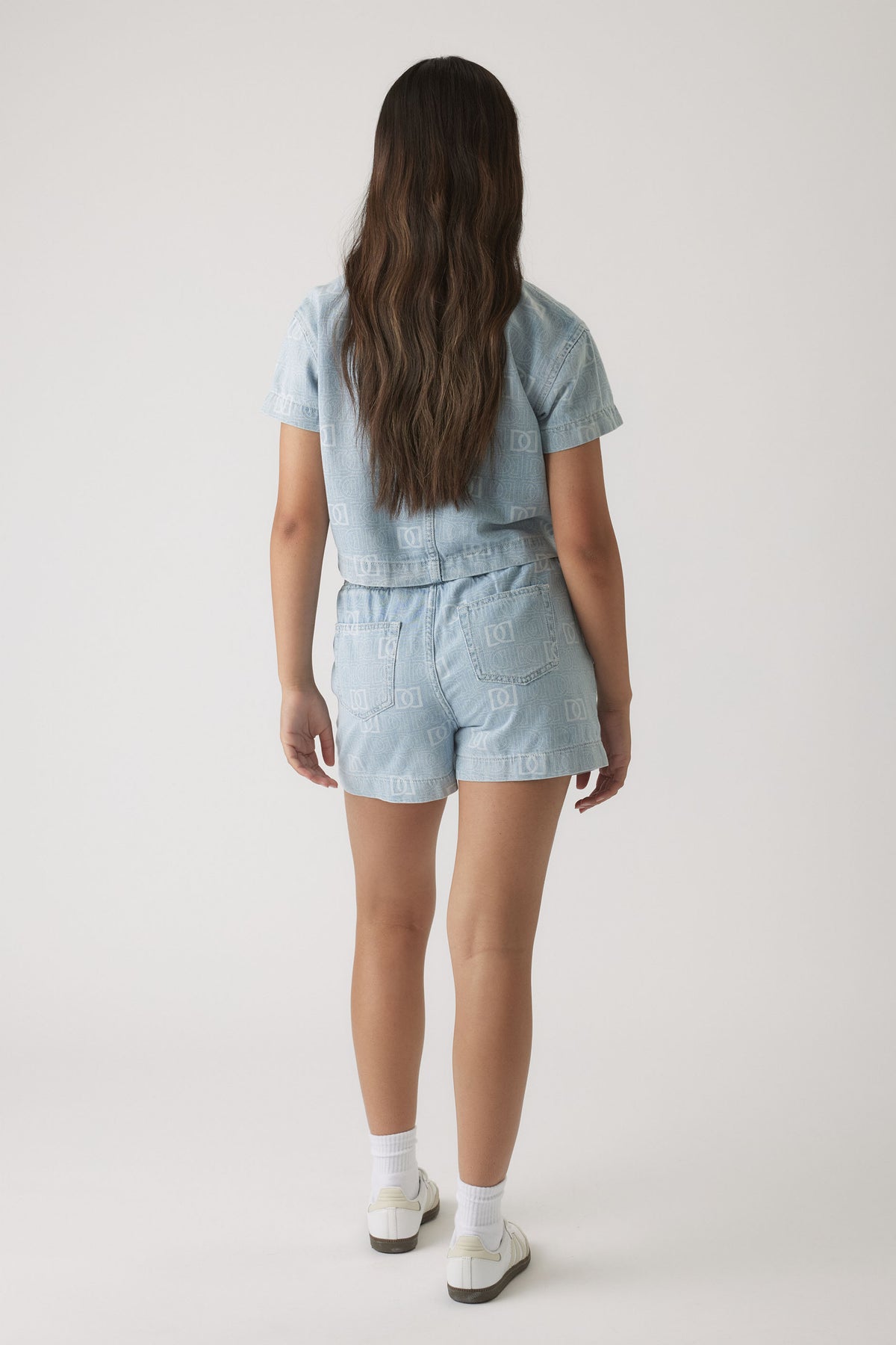 Girl wearing light blue denim shorts and matching short-sleeve shirt with subtle monogram print, paired with white sneakers.