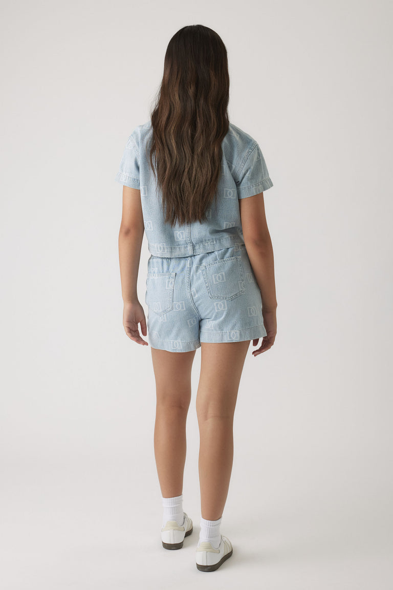 Girl wearing light blue denim shorts and matching short-sleeve shirt with subtle monogram print, paired with white sneakers.