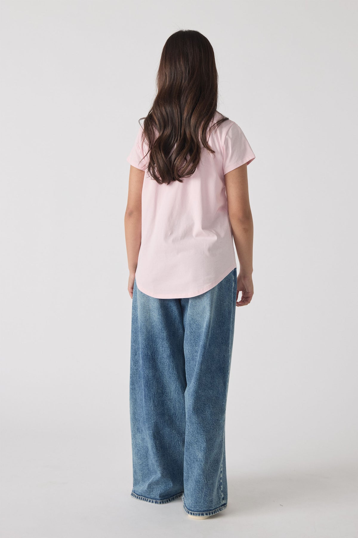 Girl shown from behind wearing a soft pink tee paired with relaxed wide-leg faded blue denim jeans.