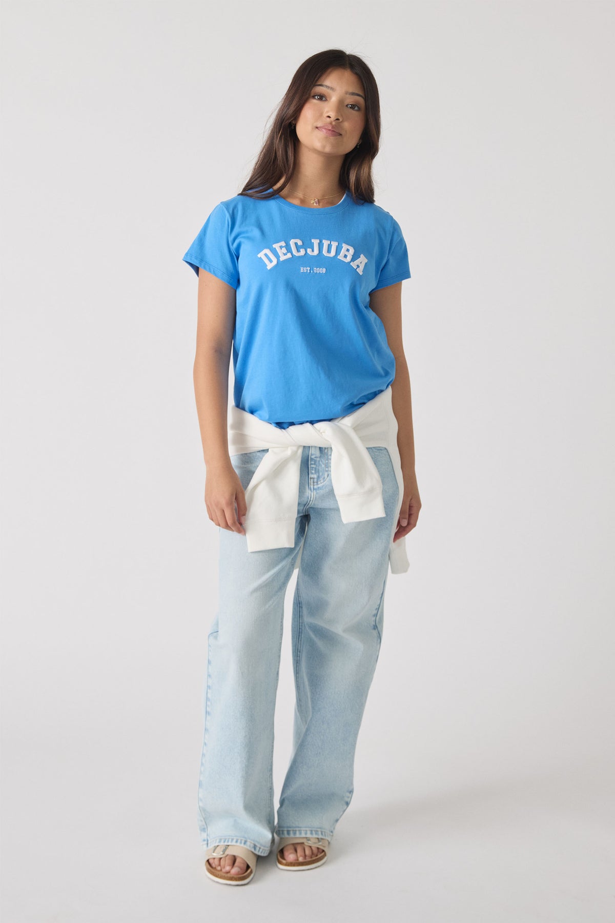 Teen girl wearing a bright blue logo tee paired with light wash wide-leg jeans and a white sweater tied at the waist.