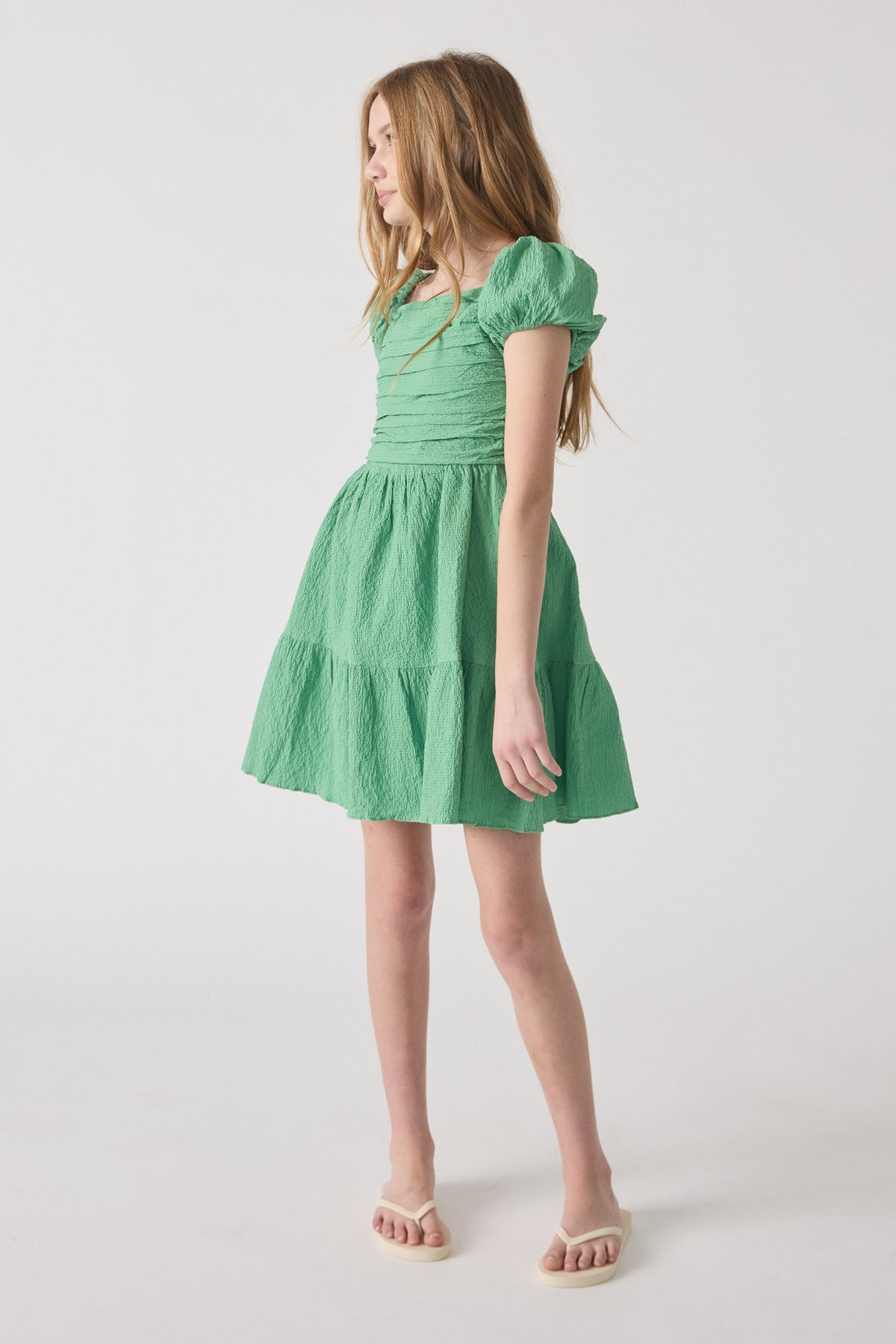 Girl wearing a spring green textured mini dress with puff sleeves and a gathered waist, paired with cream sandals