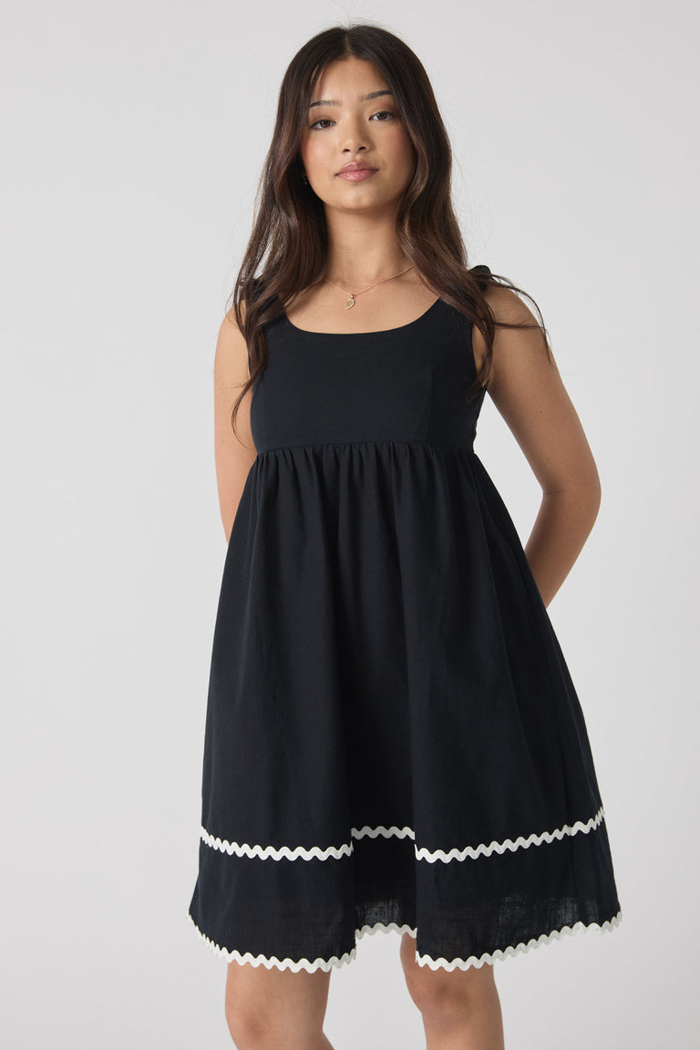 Girl wearing a sleeveless black mini dress with white wavy trim details along the hem, standing with hands behind back.