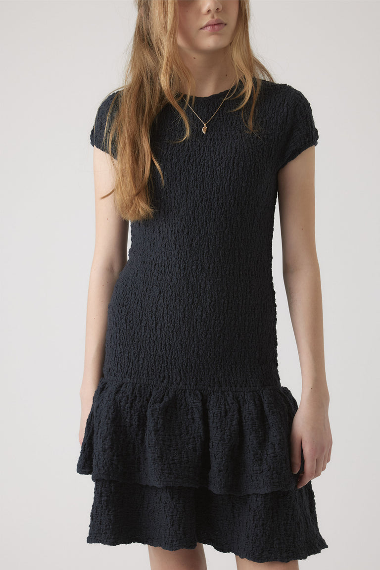 Girl wearing a textured navy shirred dress with cap sleeves and layered ruffle hem, styled with a delicate necklace.