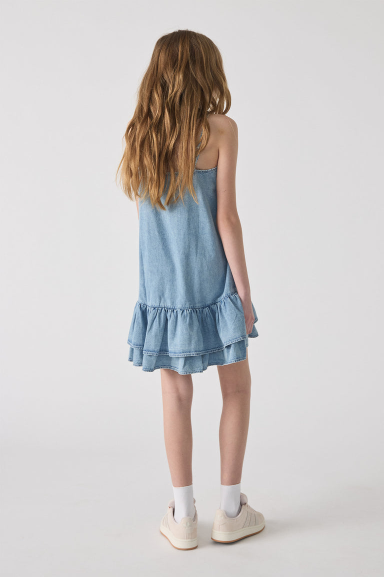 Girl seen from behind wearing a light blue denim dress with ruffle hem, white socks, and cream sneakers on plain background