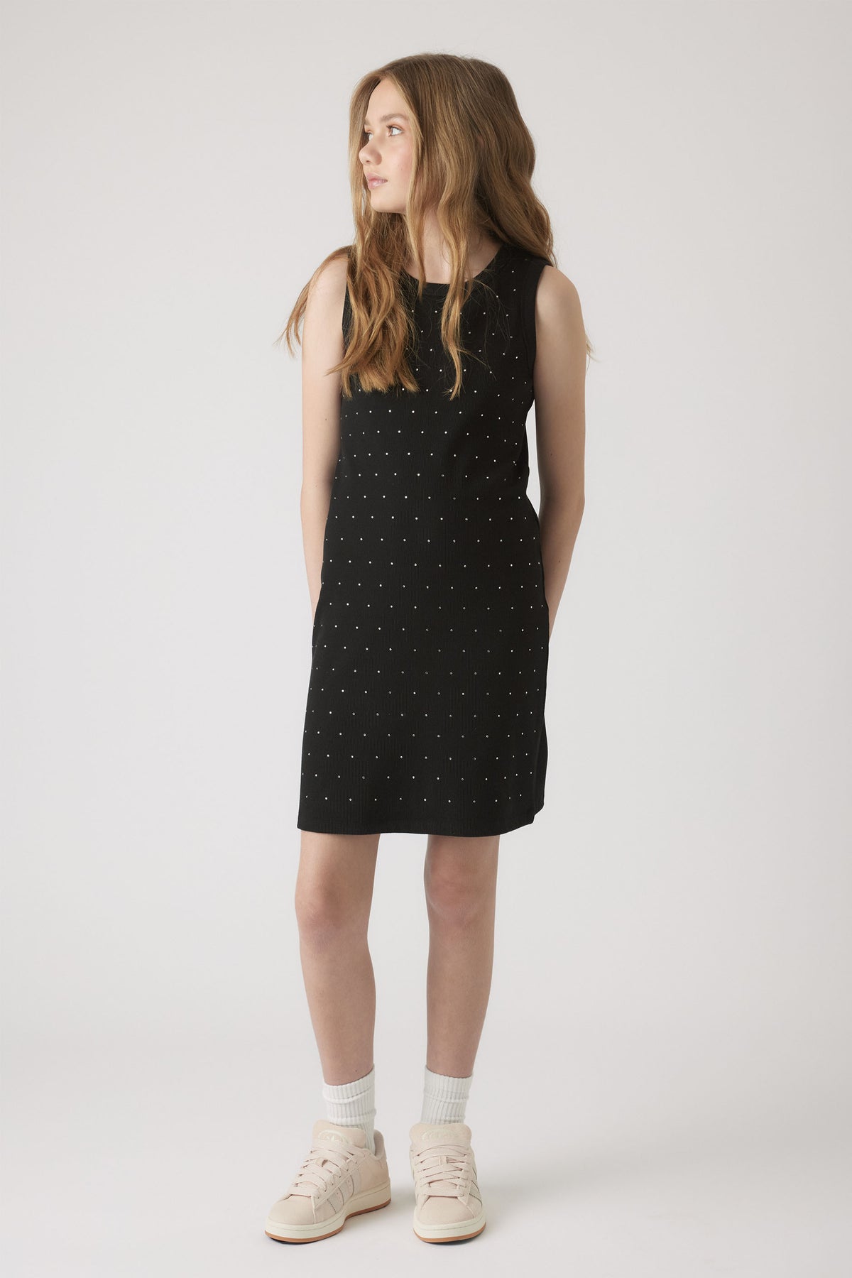 Young girl wearing a sleeveless black dress embellished with small crystals, paired with white socks and sneakers.