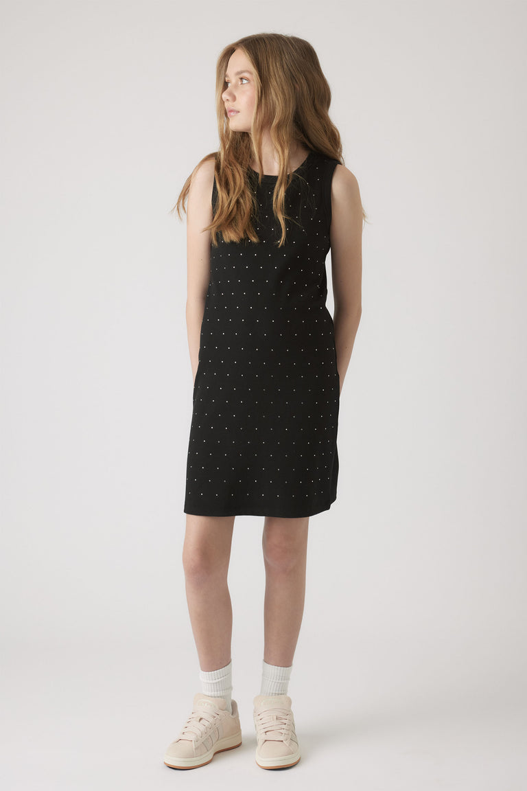 Young girl wearing a sleeveless black dress embellished with small crystals, paired with white socks and sneakers.