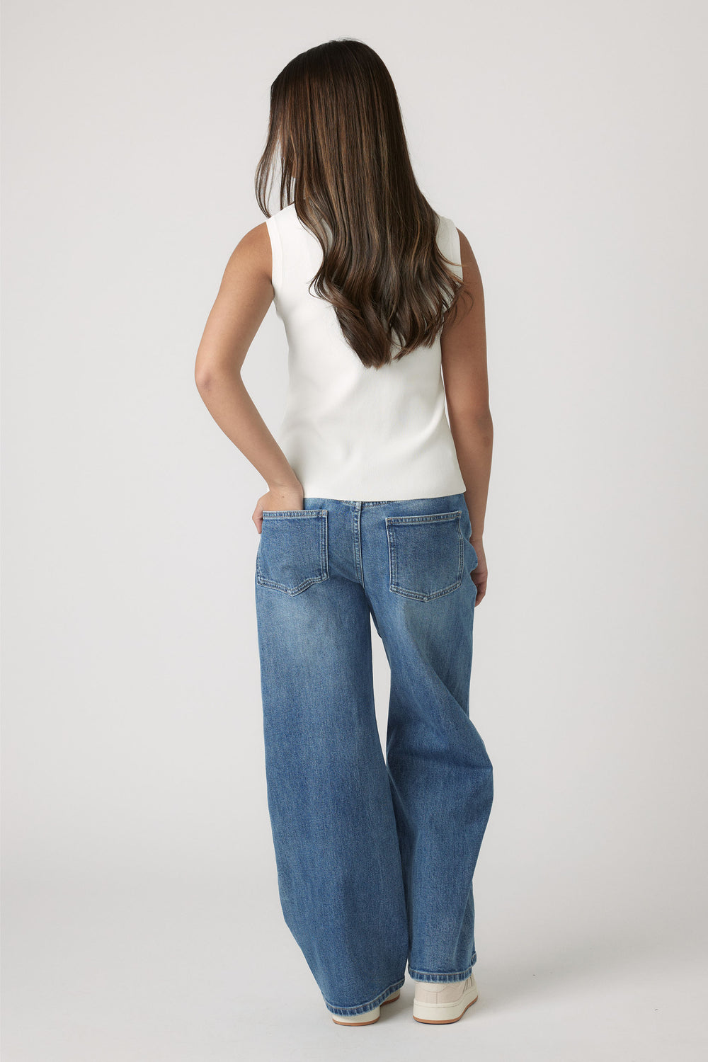 Young girl wearing a cream knit vest paired with relaxed fit blue denim jeans in a casual pose from behind