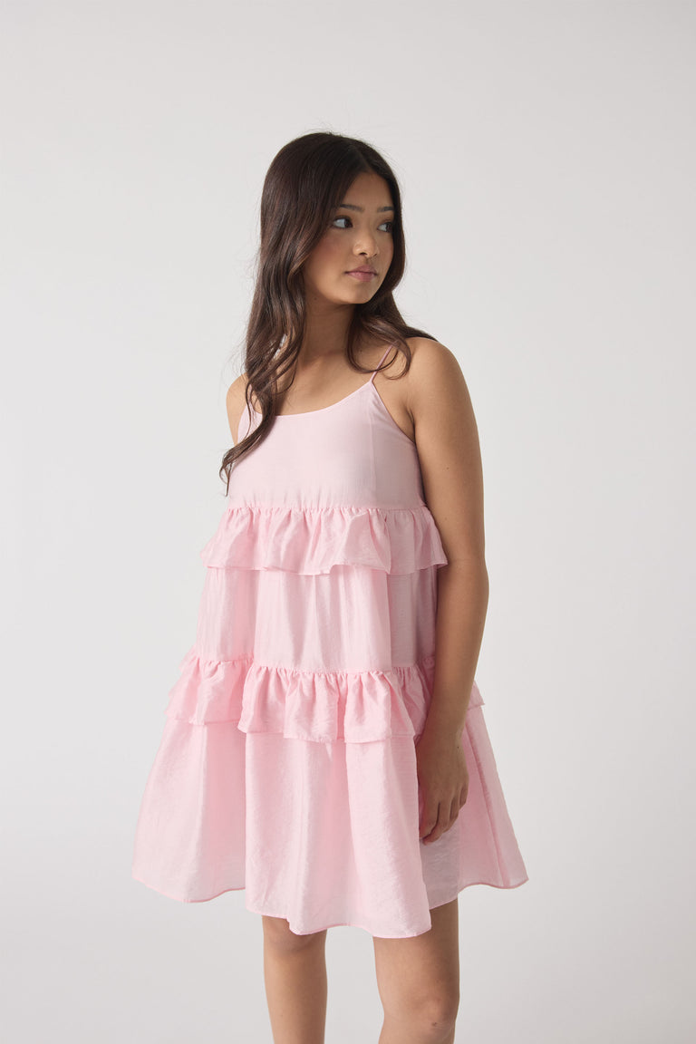 Girl wearing a soft pink tiered ruffle dress with thin straps, standing against a light neutral background.