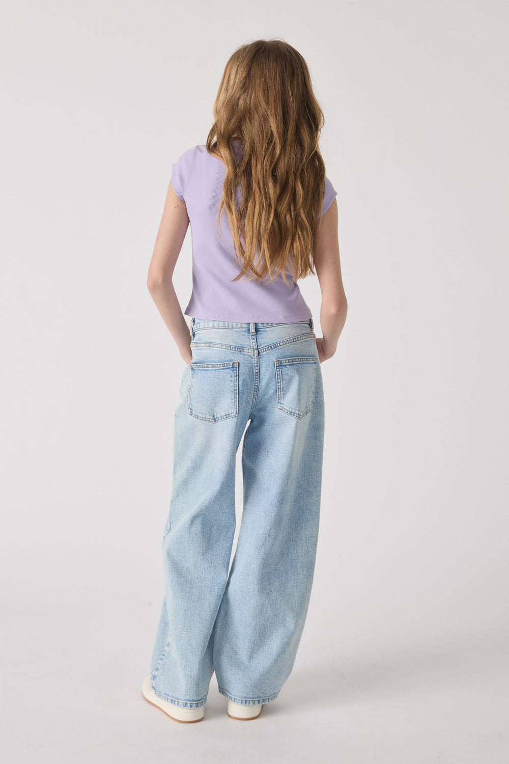 Girl wearing a lavender ribbed tee paired with light wash relaxed-fit wide-leg jeans and white sneakers against a plain backdrop.