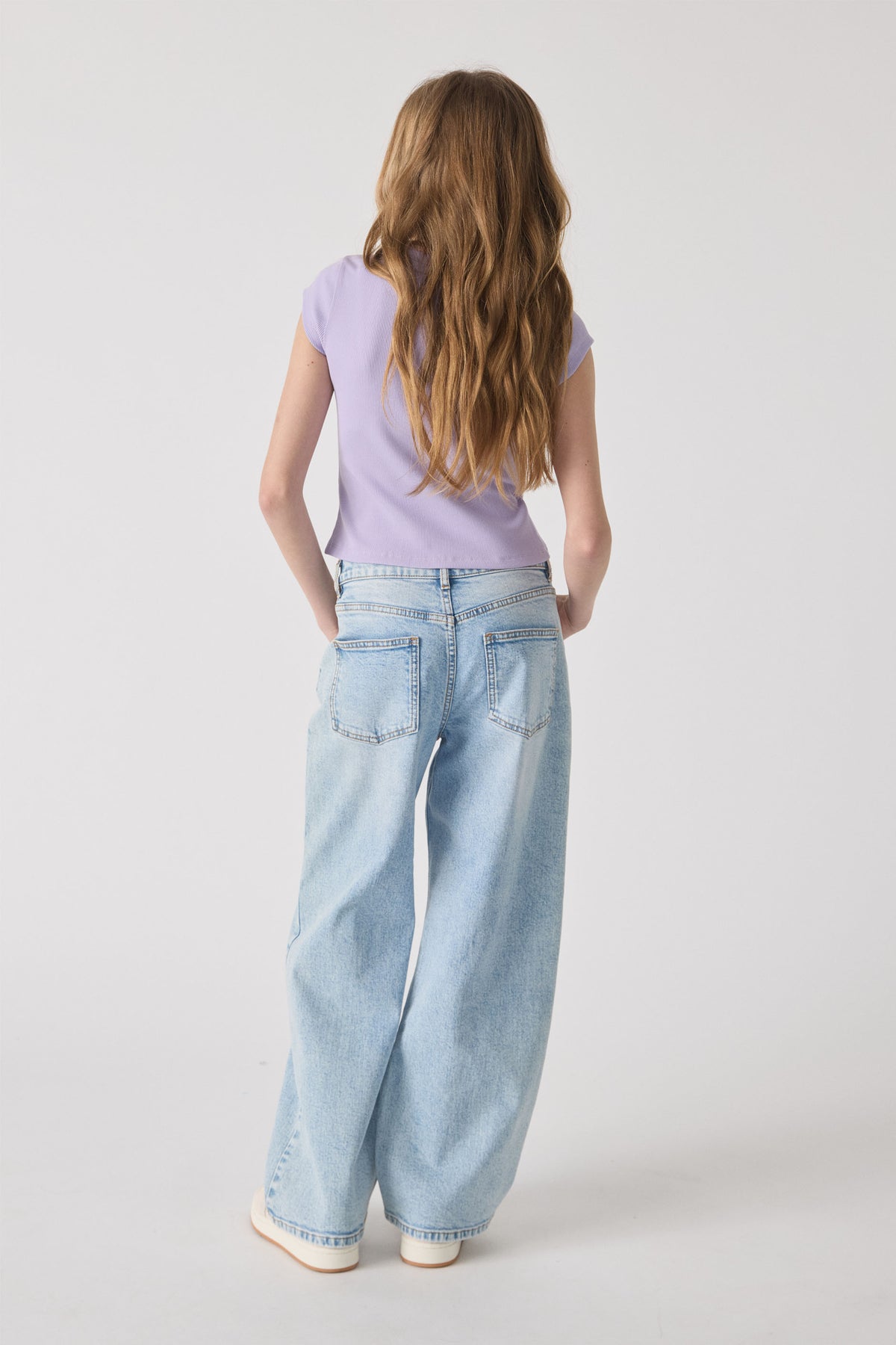 Girl wearing a lavender ribbed tee paired with light wash relaxed-fit wide-leg jeans and white sneakers against a plain backdrop.