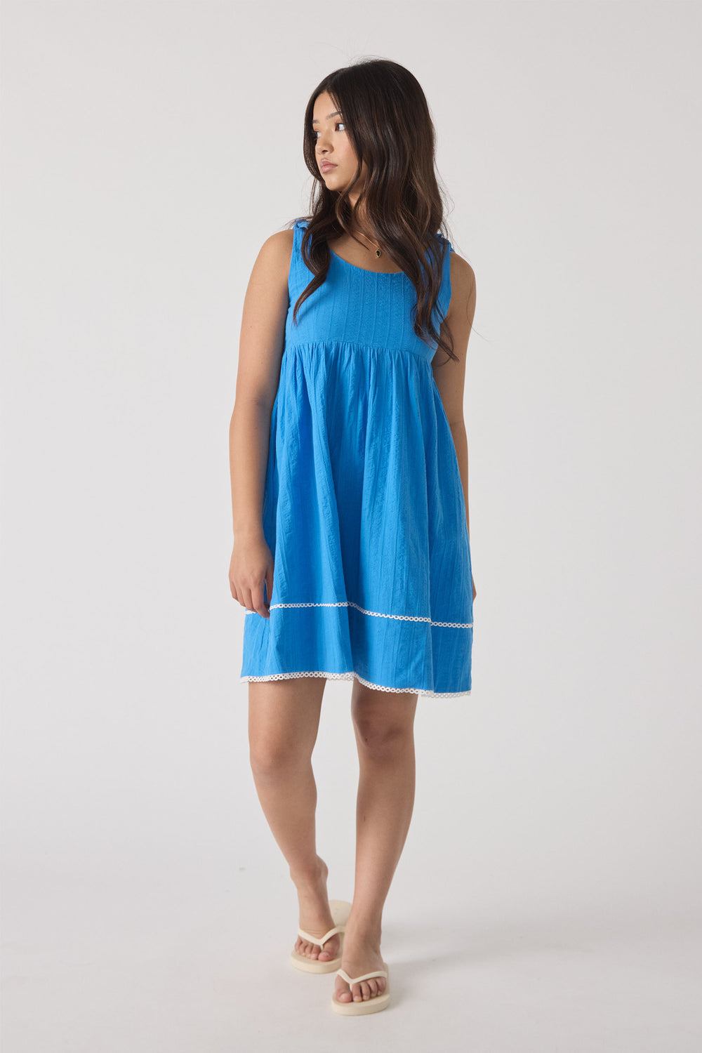 Girl wearing a bright blue sleeveless mini dress with subtle textured details and white trim, paired with beige sandals.