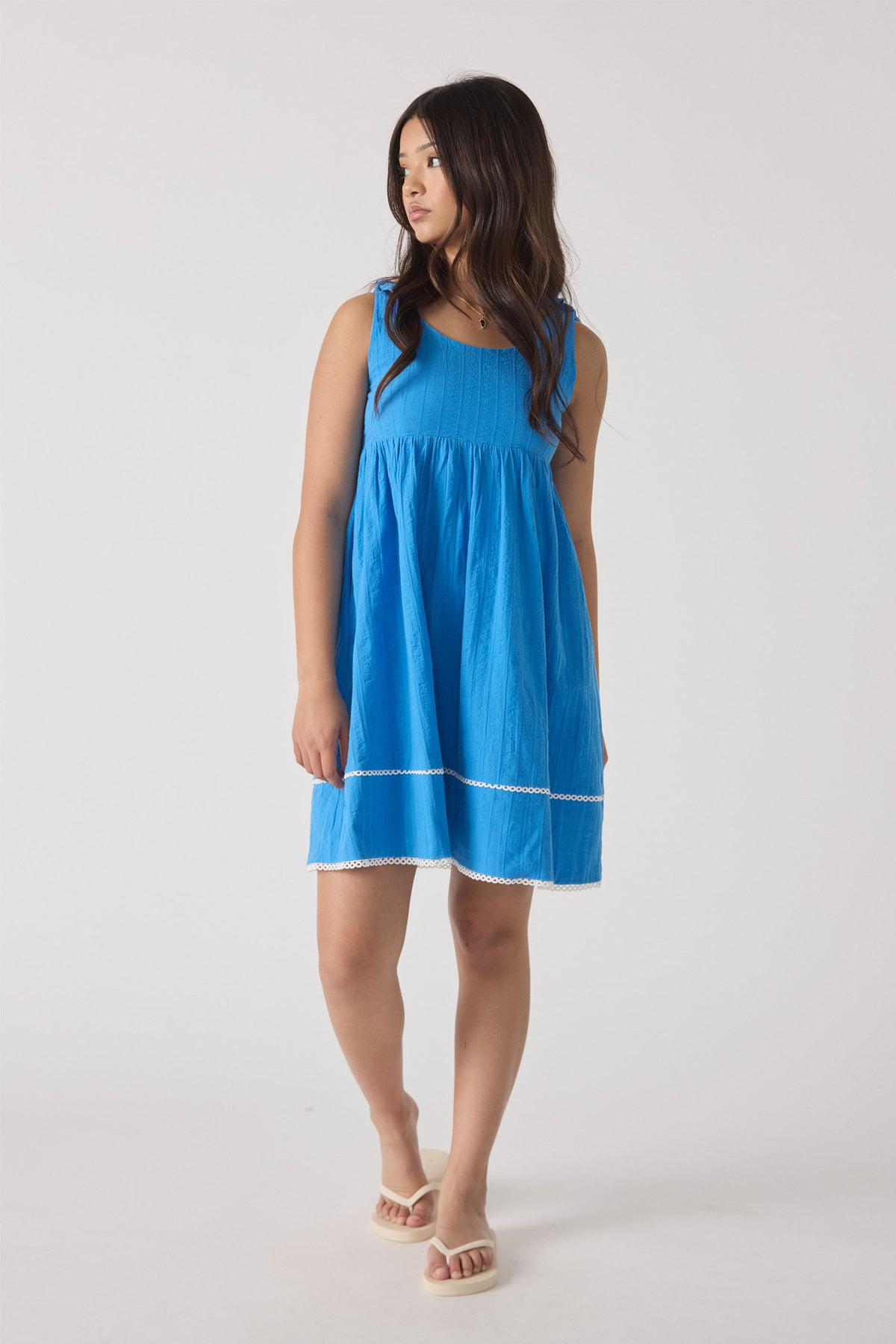 Girl wearing a bright blue sleeveless mini dress with subtle textured details and white trim, paired with beige sandals.