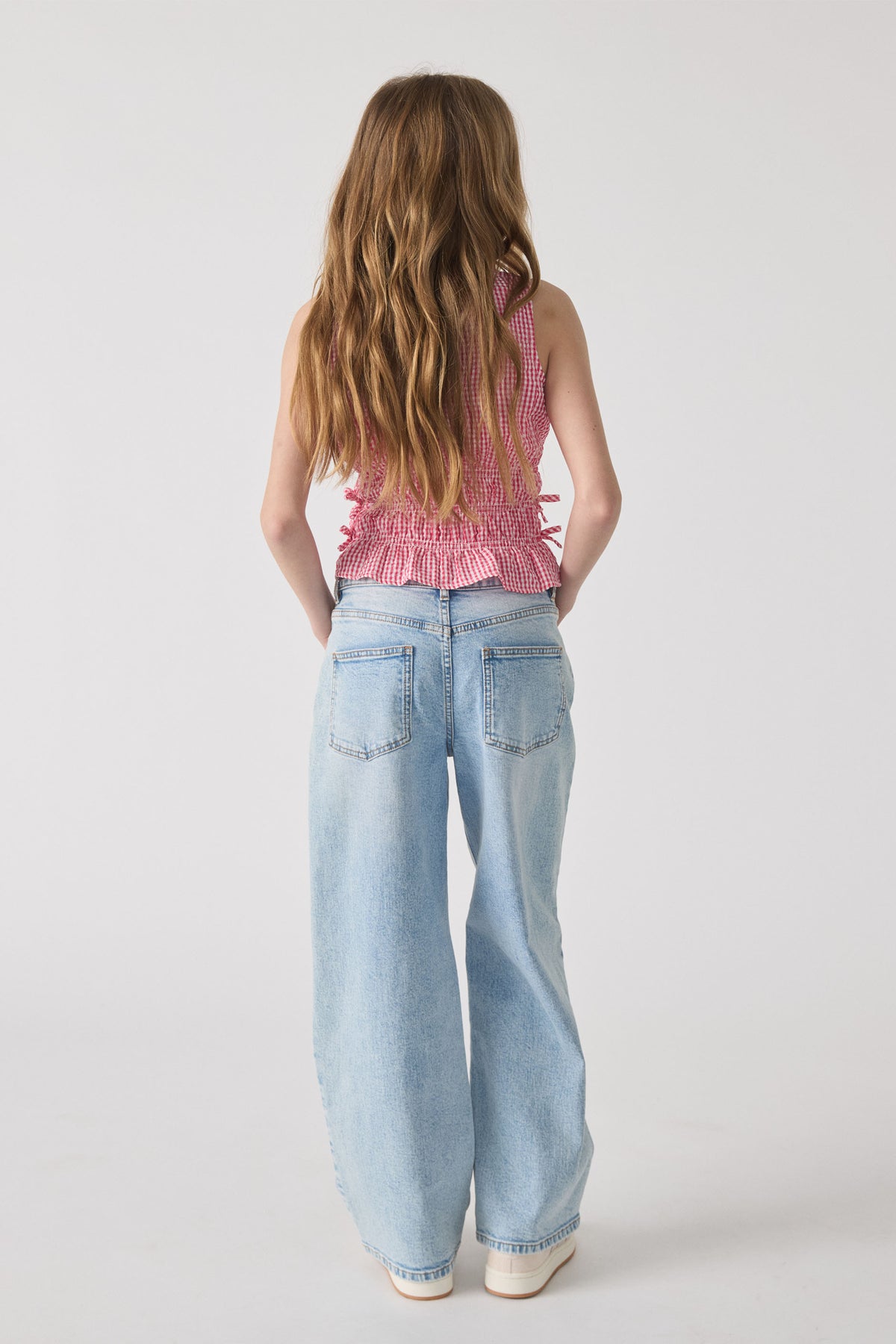 Girl shown from behind wearing a sleeveless red gingham tie top paired with light wash wide-leg denim jeans and white shoes