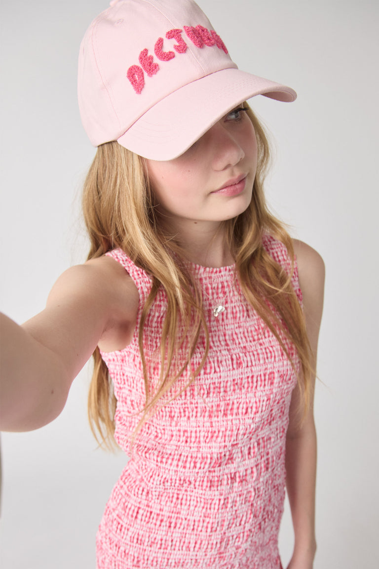 Young girl wearing a textured pink sleeveless dress paired with a soft pink cap featuring bold embroidery.