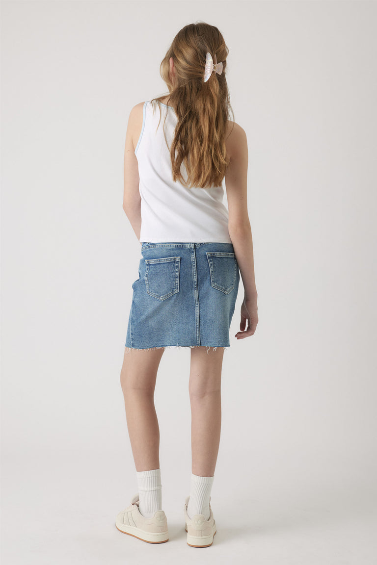 Girl wearing a white sleeveless tank paired with a frayed hem denim skirt, white socks, and cream sneakers.