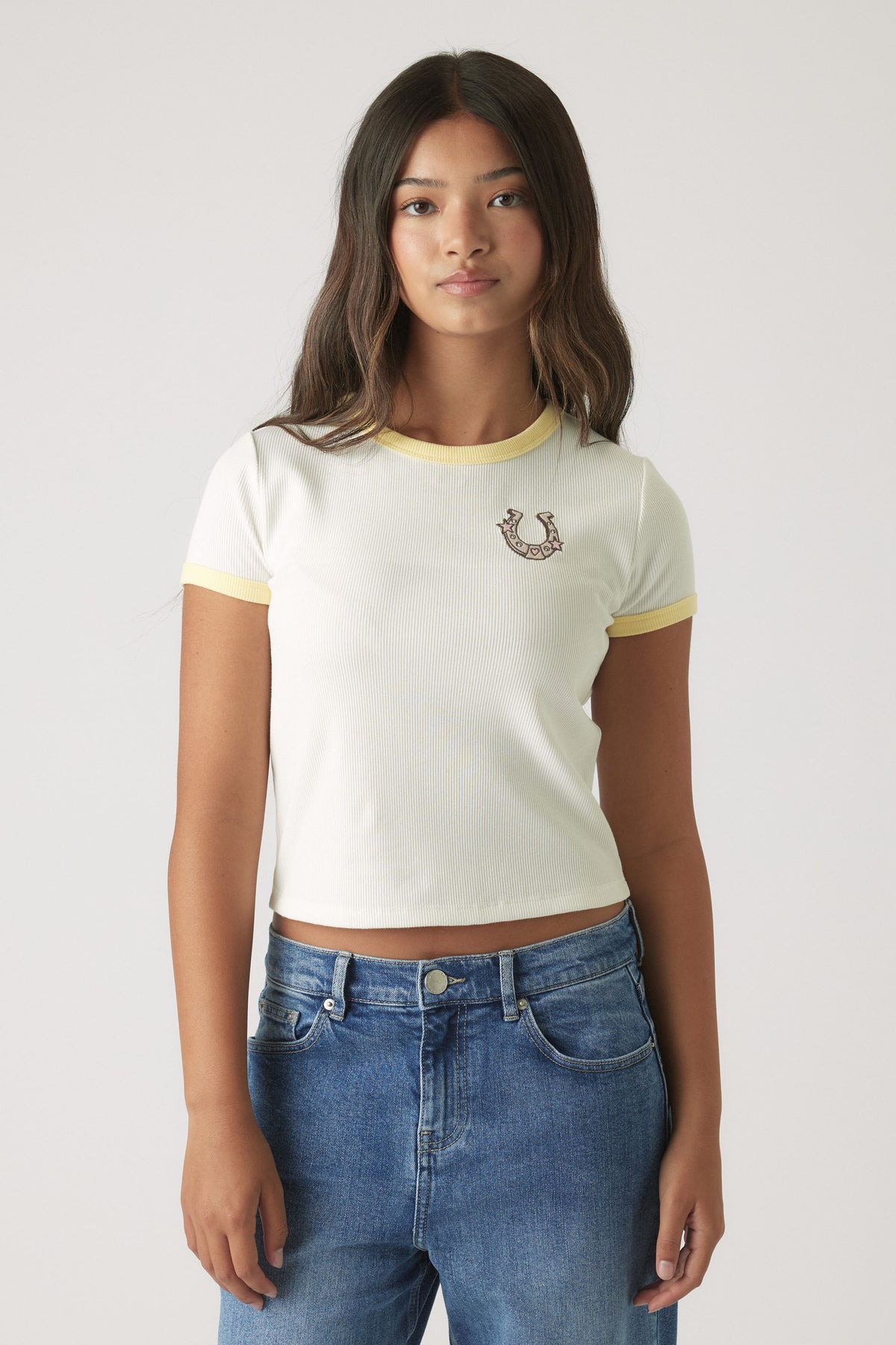 Girl wearing a white cropped ringer tee with yellow trim and embroidered design paired with relaxed blue denim jeans