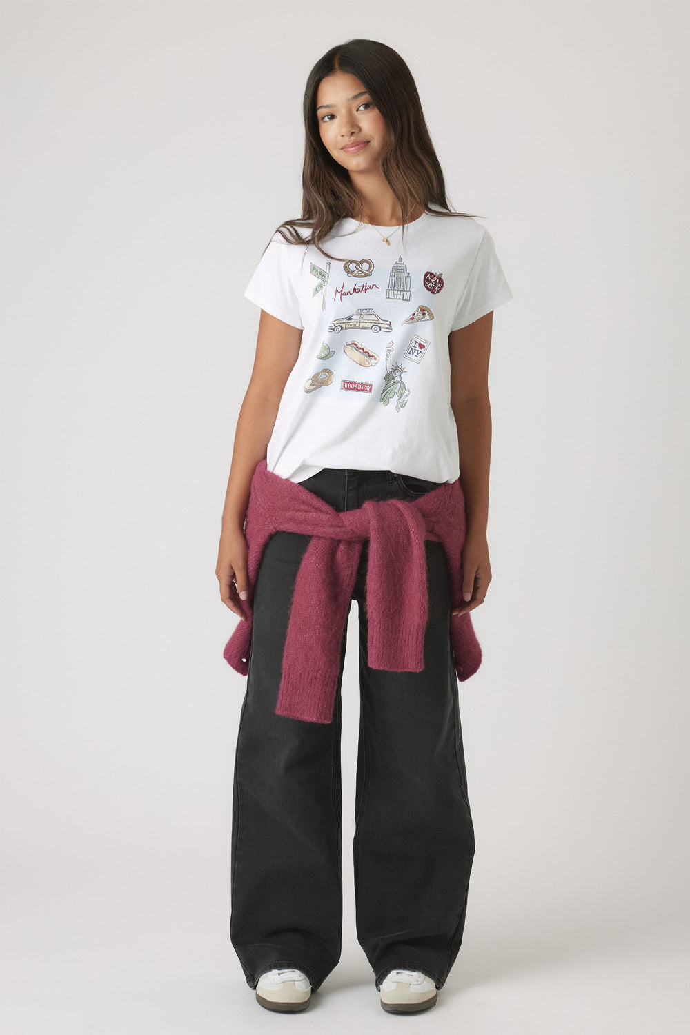 Portrait of a girl wearing Girls New York City Tee with City-themed embellishments, styled with black pants and a tied sweater.