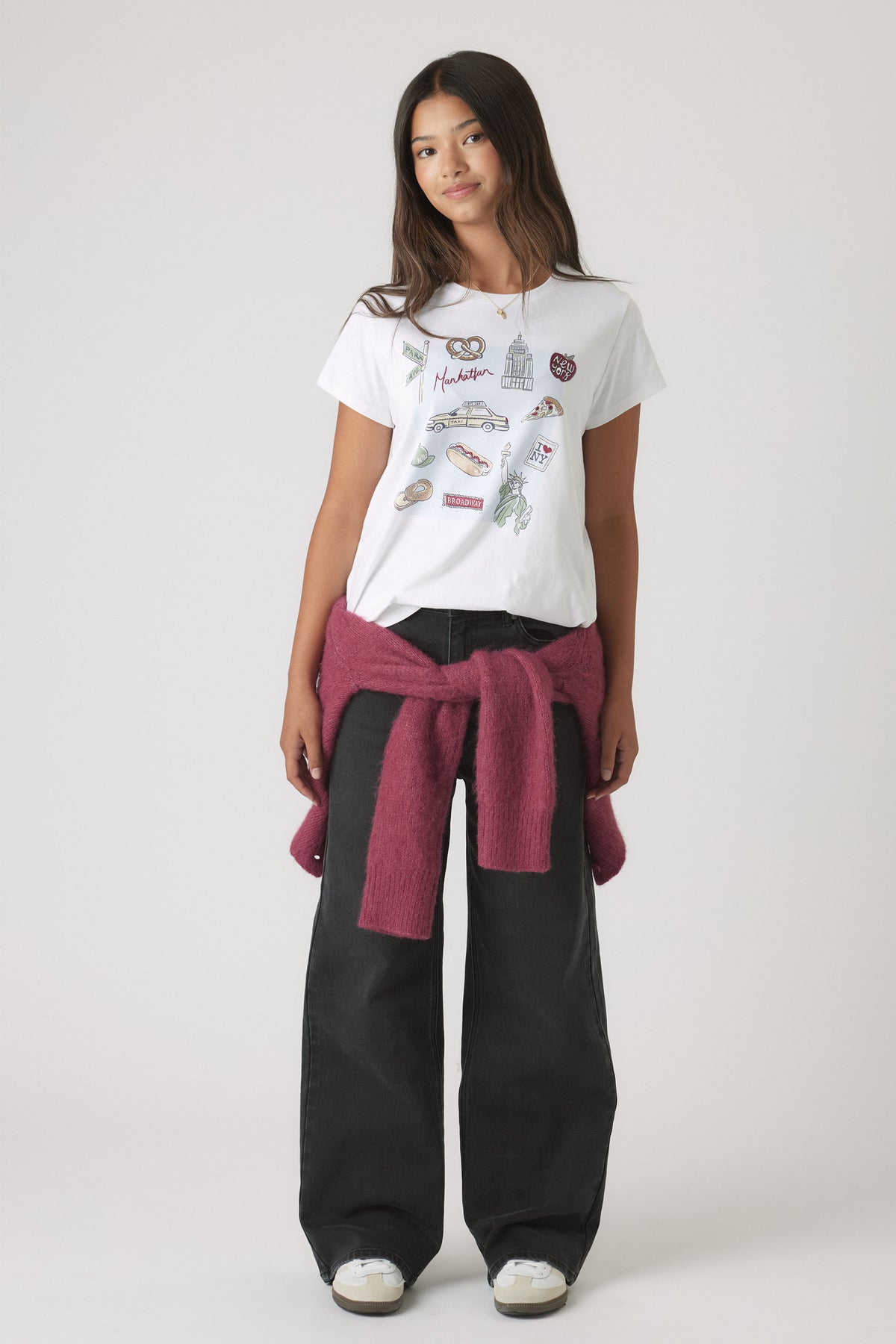 Portrait of a girl wearing Girls New York City Tee with City-themed embellishments, styled with black pants and a tied sweater.