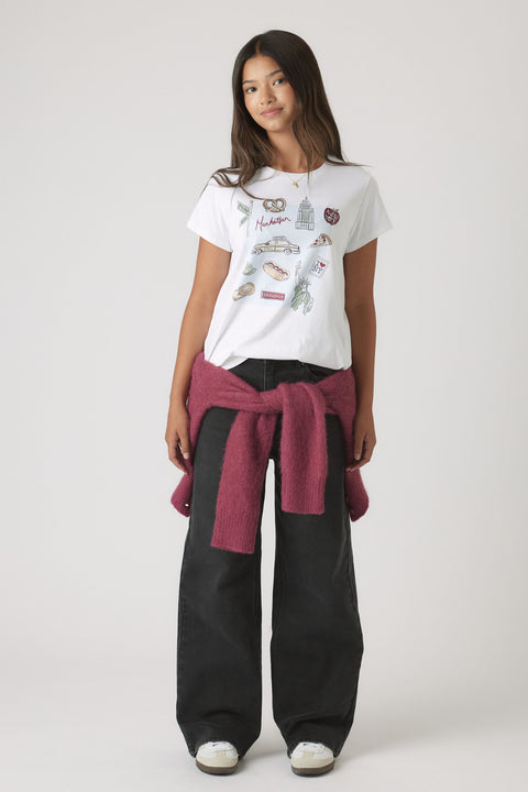 Portrait of a girl wearing Girls New York City Tee with City-themed embellishments, styled with black pants and a tied sweater.