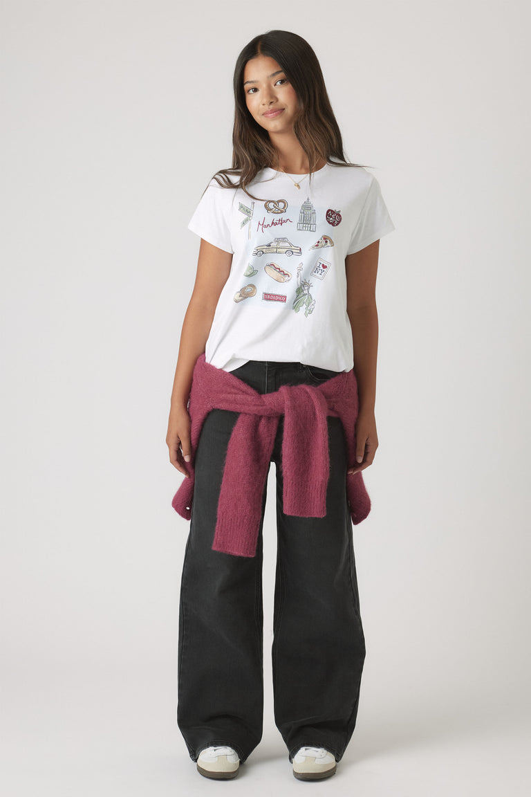 Portrait of a girl wearing Girls New York City Tee with City-themed embellishments, styled with black pants and a tied sweater.
