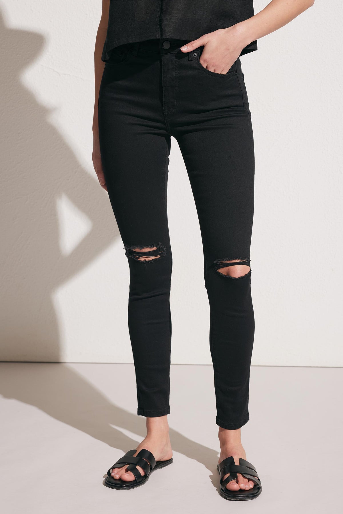 Woman wearing black distressed skinny jeans paired with black slide sandals against a neutral background
