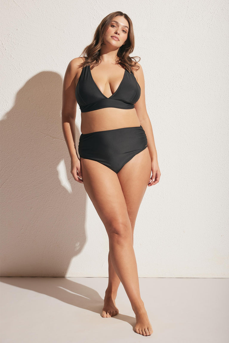 Plus-size woman wearing a black matte triangle bikini top and high-waisted bikini bottoms against a light wall background