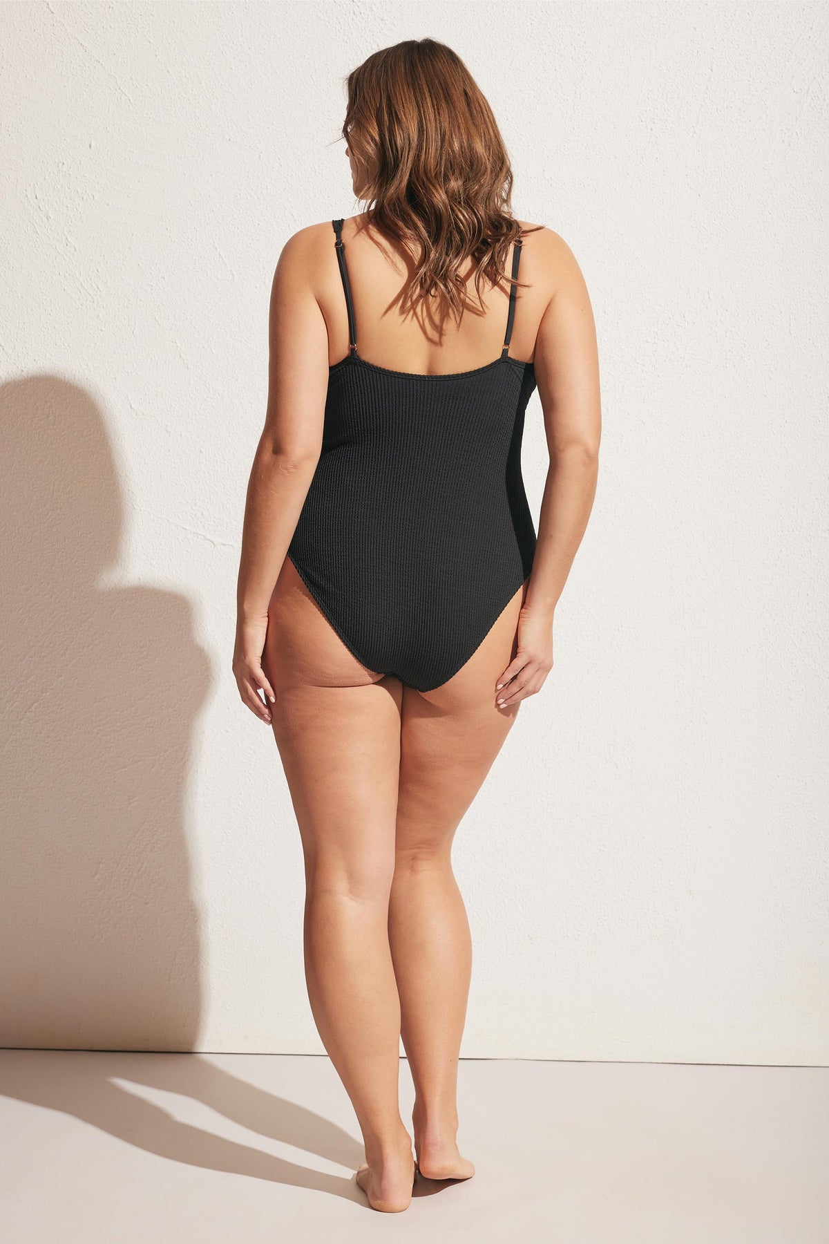 Woman wearing a black ribbed one-piece with adjustable shoulder straps, shown from the back against a light wall.
