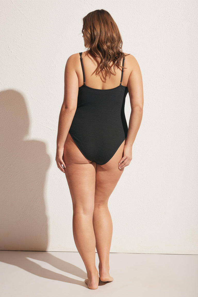 Woman wearing a black ribbed one-piece with adjustable shoulder straps, shown from the back against a light wall.