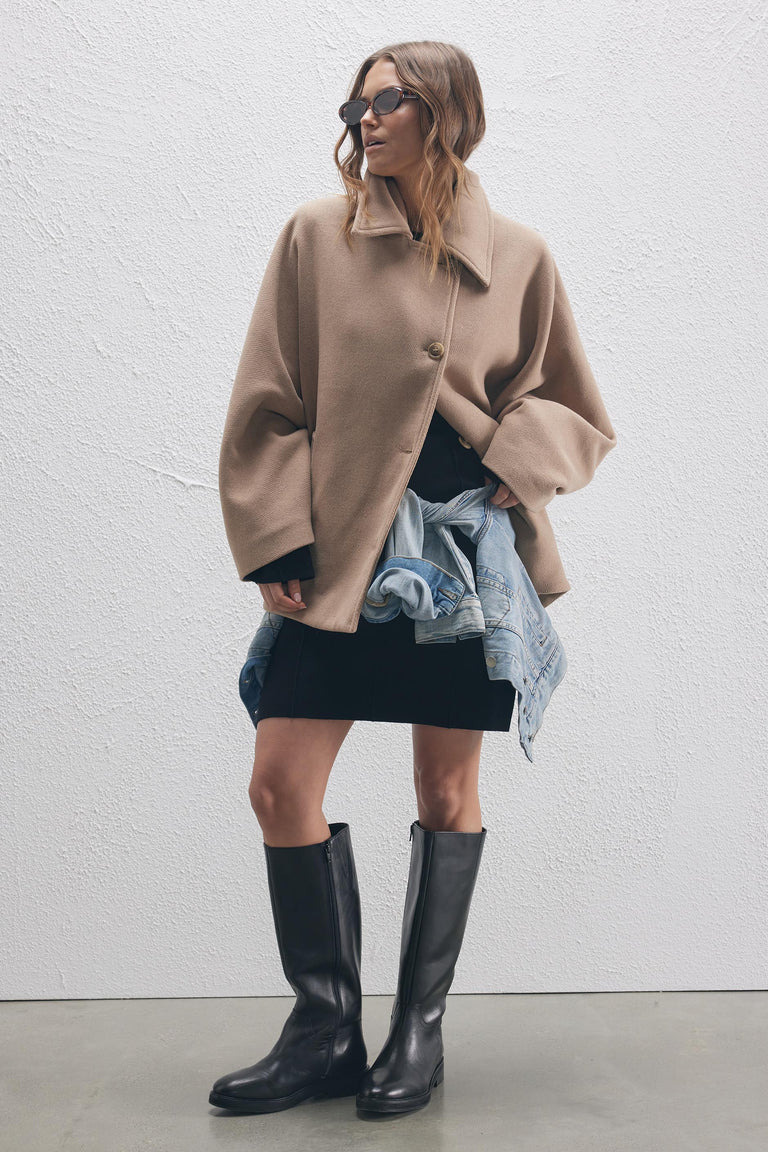 Woman wearing oval tortoiseshell sunglasses with a tan oversized coat, black dress, denim jacket tied at waist, and black boots