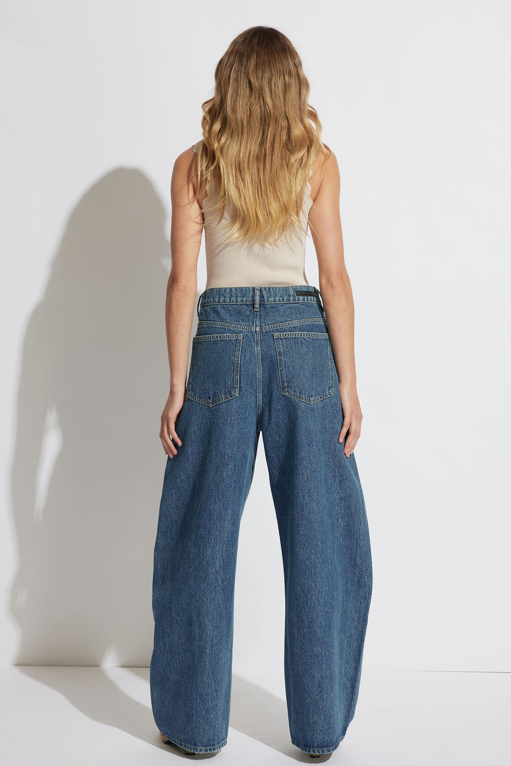 Woman with long wavy blonde hair wearing a beige scallop edge tank and wide-leg blue denim jeans standing against a white wall