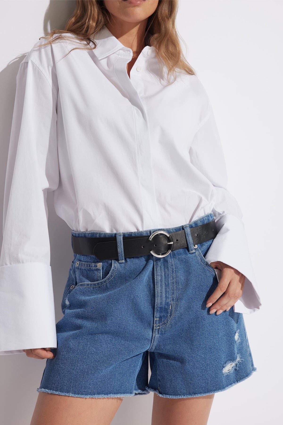 Woman wearing mid-blue distressed denim shorts with raw hems and a black belt, paired with a white button-up shirt