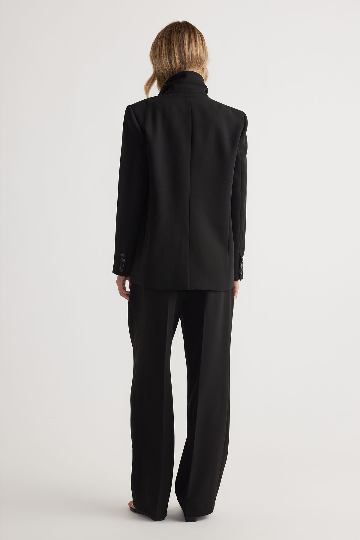 Back view of woman wearing black tailored blazer with structured shoulders and matching wide-leg pants against white background