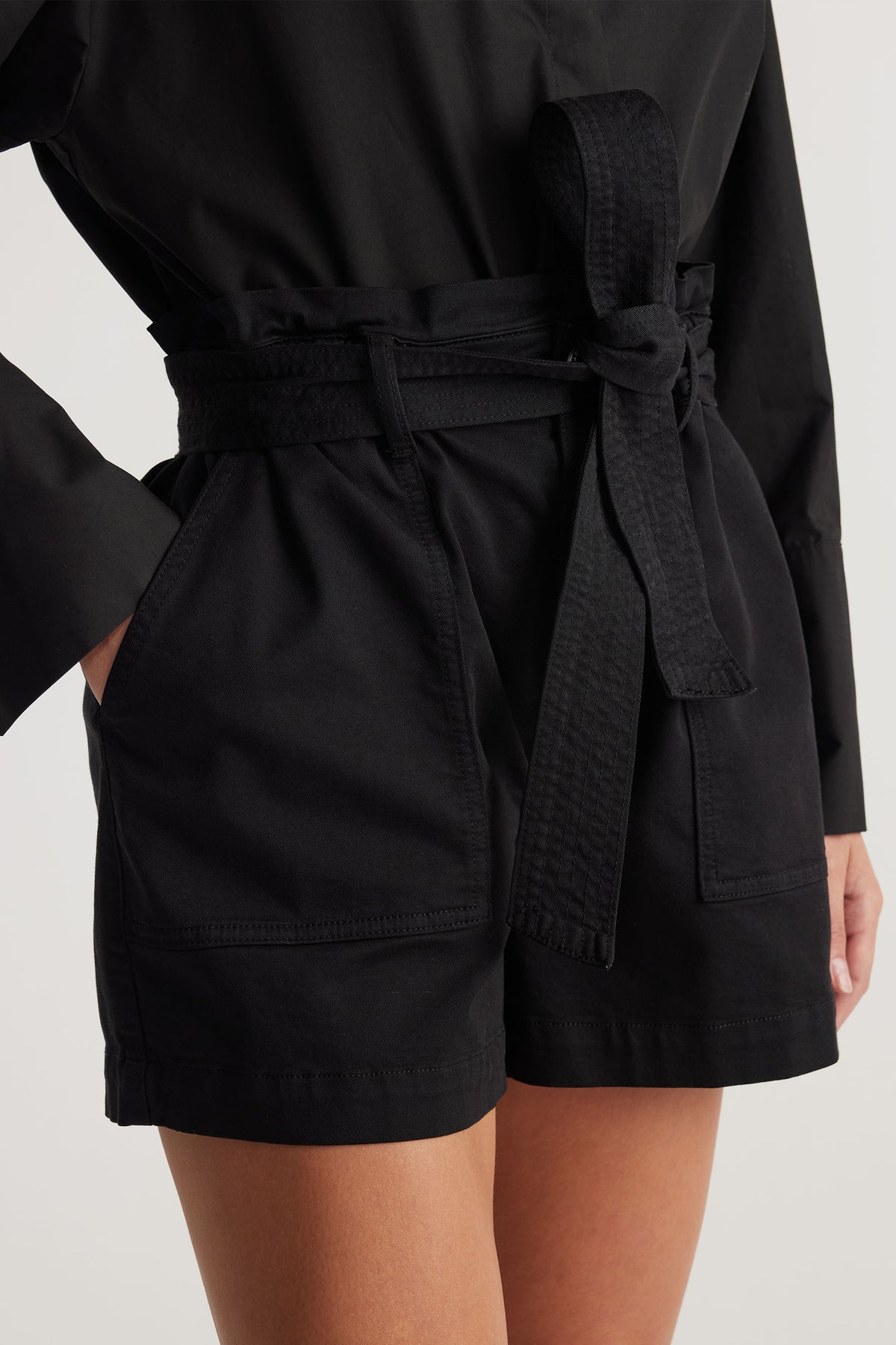 Close-up of woman wearing black high-waisted shorts with a tied belt and large front pockets styled with a long-sleeve top