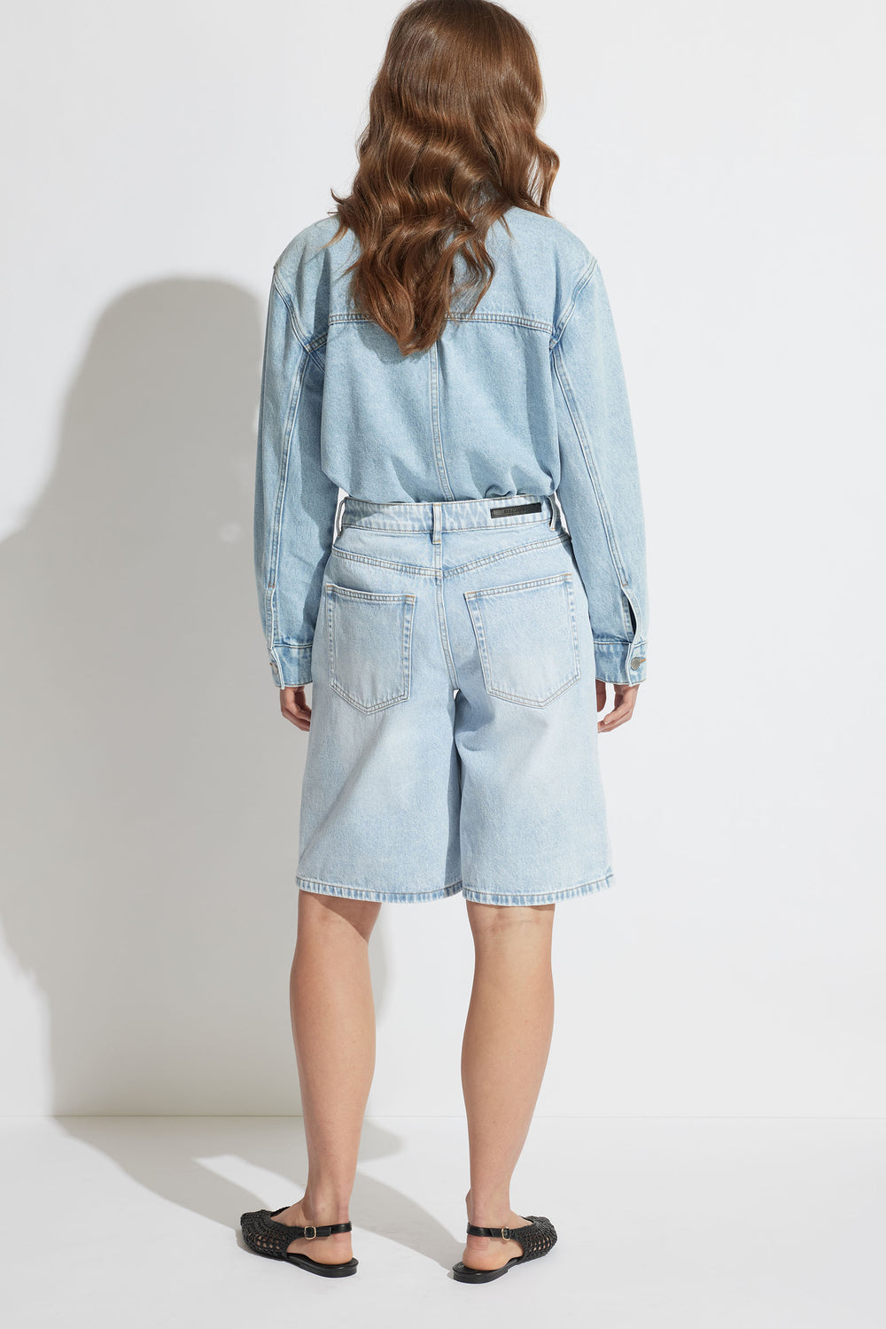 Woman seen from behind wearing light blue longline denim shorts paired with matching denim jacket and black sandals