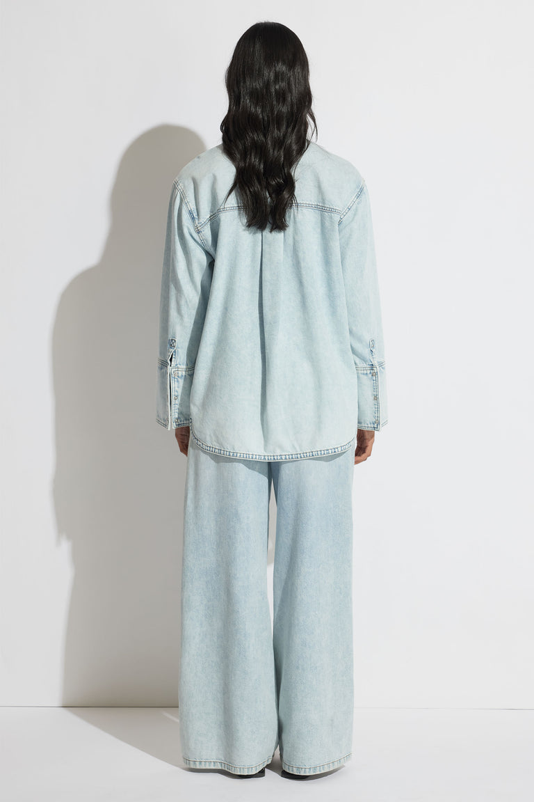 Woman wearing a relaxed fit light blue denim shirt and wide-leg denim pants, shown from the back against a plain wall.