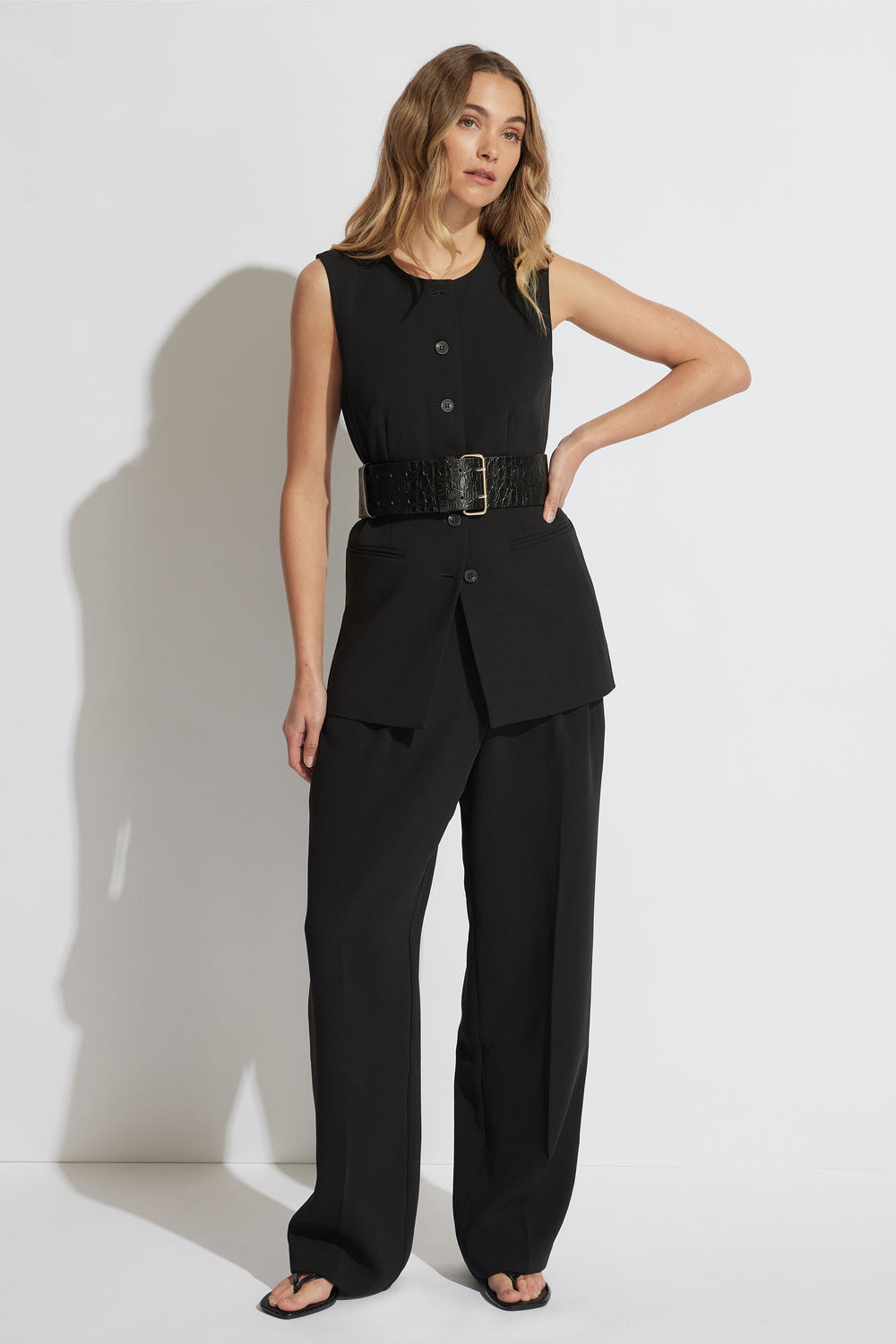 Woman wearing a sleeveless black tailored vest with a wide textured belt and matching wide-leg pants.