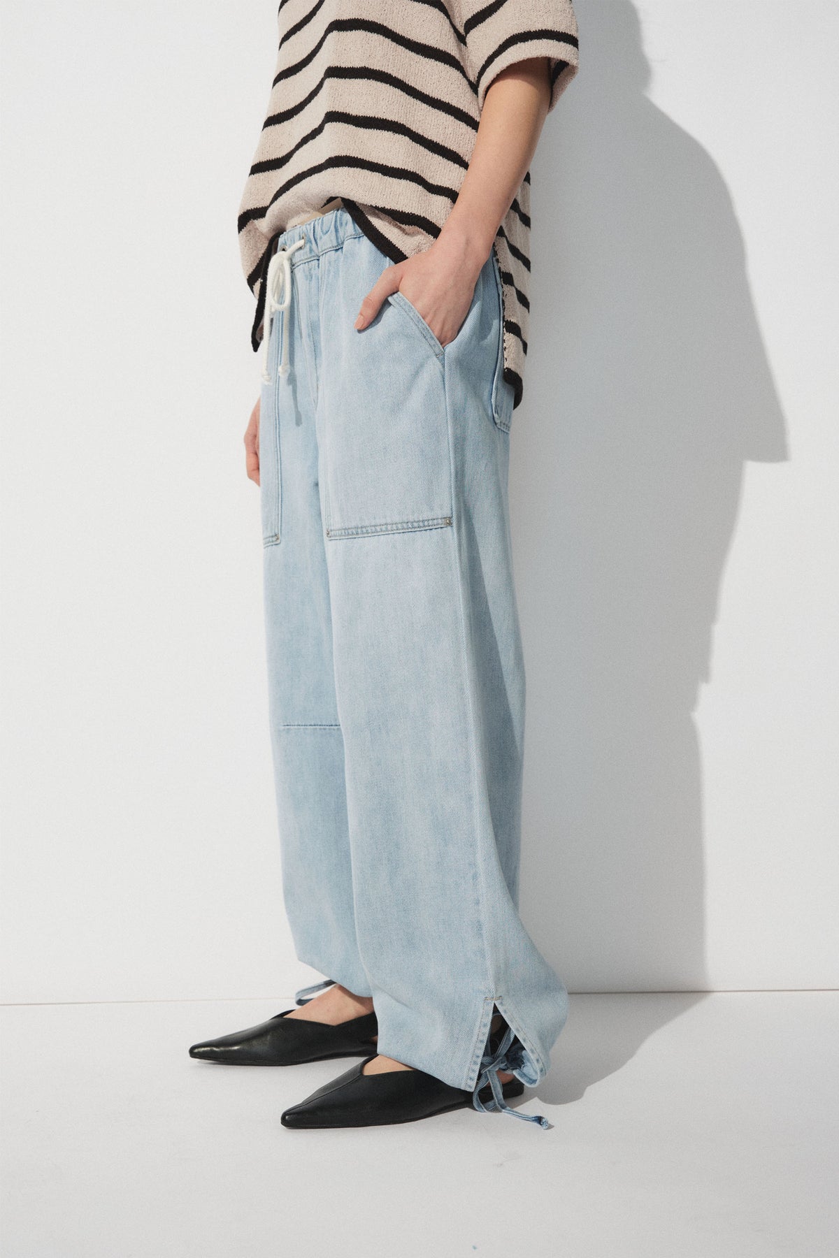 Side view of light blue wide-leg denim joggers with drawstring waist and ankle ties paired with black flats and striped top
