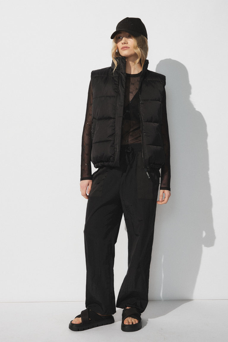 Woman wearing a black puffer vest over a sheer long-sleeve top with loose black joggers and sandals, black cap.