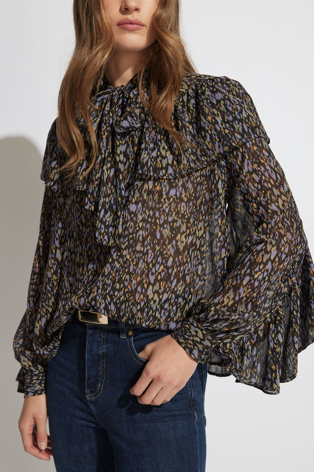 Woman wearing a sheer patterned blouse with ruffle details and long puffed sleeves paired with dark denim jeans.