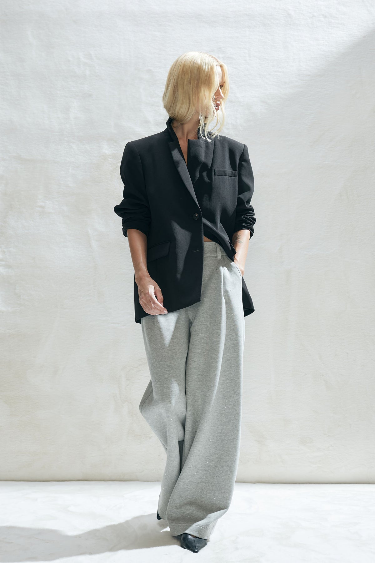 Woman wearing a relaxed black blazer over loose light grey pull-on pants with hands in pockets, standing against a bright backdrop.