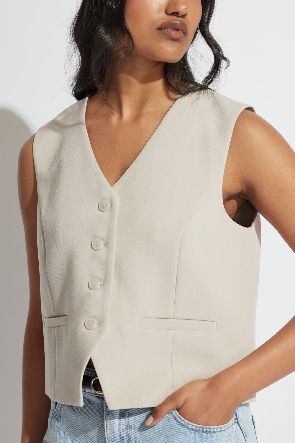 Woman wearing a sleeveless tailored vest in soft beige with button front and welt pockets, styled with light denim jeans.