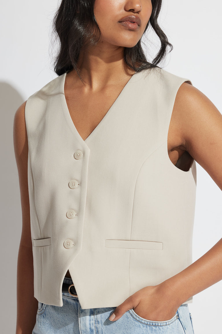 Woman wearing a sleeveless tailored vest in soft beige with button front and welt pockets, styled with light denim jeans.